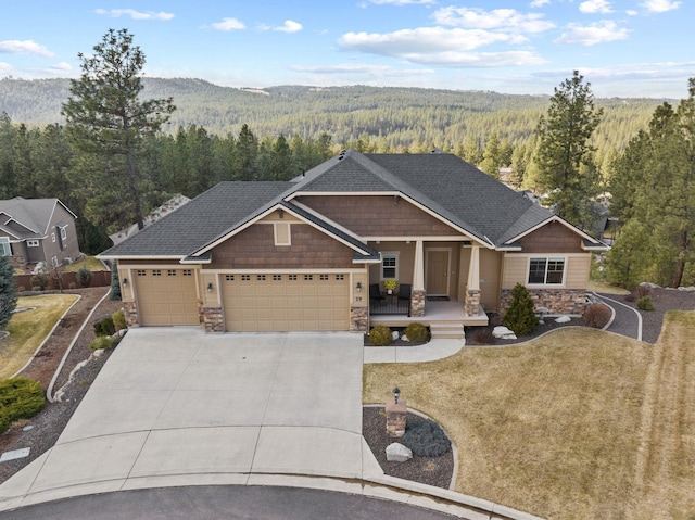 craftsman house with covered porch, a garage, stone siding, driveway, and a forest view