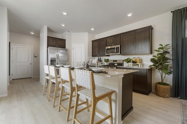 kitchen featuring appliances with stainless steel finishes, a kitchen island with sink, dark brown cabinets, light wood-type flooring, and a kitchen breakfast bar