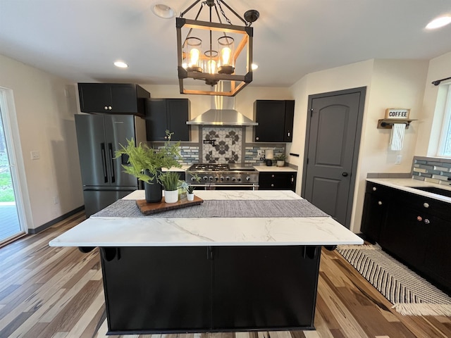 kitchen with appliances with stainless steel finishes, tasteful backsplash, light wood-type flooring, dark cabinetry, and wall chimney exhaust hood