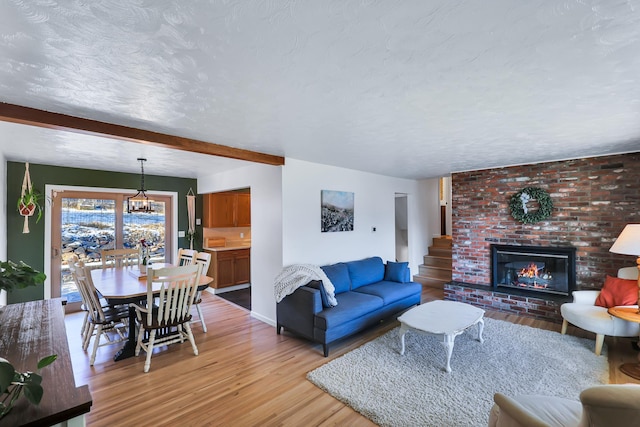 living room with beam ceiling, light wood-style flooring, an inviting chandelier, a brick fireplace, and a textured ceiling