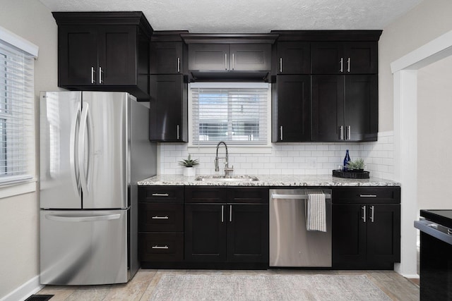 kitchen with light stone counters, a sink, appliances with stainless steel finishes, dark cabinetry, and tasteful backsplash