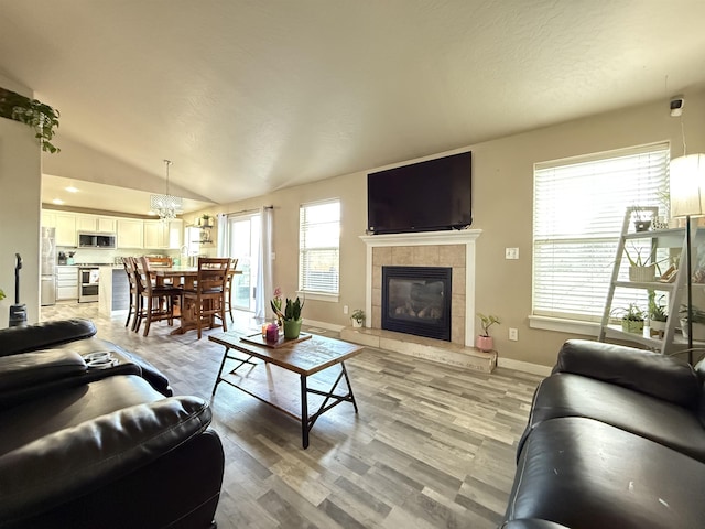 living area featuring baseboards, a tiled fireplace, lofted ceiling, an inviting chandelier, and light wood-type flooring