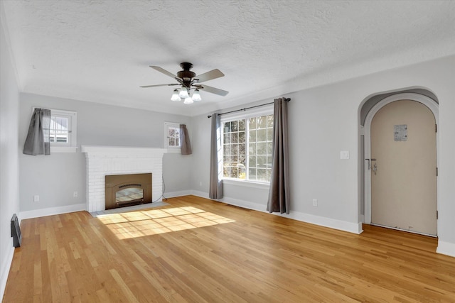 unfurnished living room with arched walkways, a fireplace, a ceiling fan, a textured ceiling, and light wood-type flooring