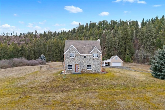 view of front facade with a garage, a forest view, stone siding, an outbuilding, and a front yard