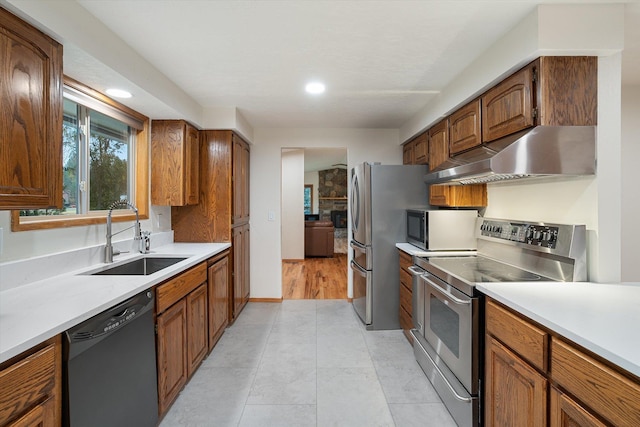 kitchen with brown cabinetry, stainless steel appliances, light countertops, under cabinet range hood, and a sink