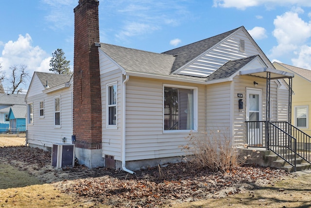 bungalow-style house with central air condition unit, a shingled roof, a chimney, and fence