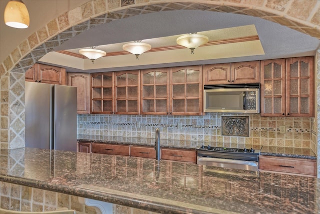 kitchen featuring appliances with stainless steel finishes, backsplash, brown cabinets, dark stone counters, and a tray ceiling