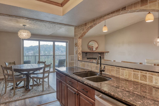 kitchen with a textured ceiling, a sink, hanging light fixtures, stainless steel dishwasher, and dark stone countertops