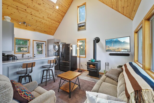 living room with a skylight, stacked washer / dryer, wood ceiling, a wood stove, and high vaulted ceiling