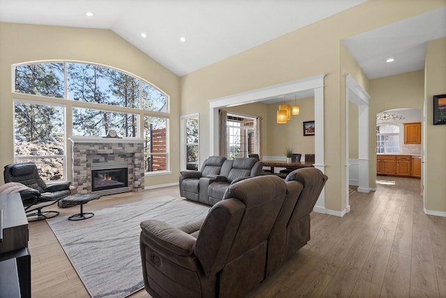 living room featuring arched walkways, high vaulted ceiling, a fireplace, baseboards, and light wood finished floors