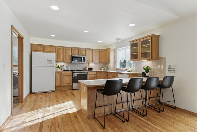 kitchen featuring light wood finished floors, decorative backsplash, glass insert cabinets, a peninsula, and stainless steel appliances