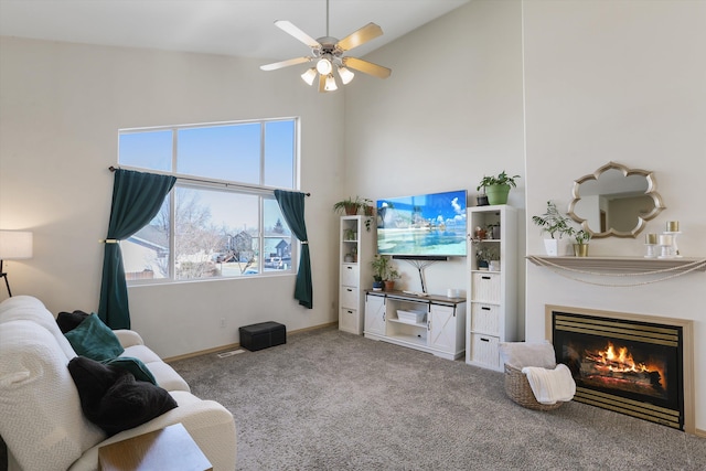 carpeted living room featuring a warm lit fireplace, ceiling fan, a towering ceiling, and baseboards