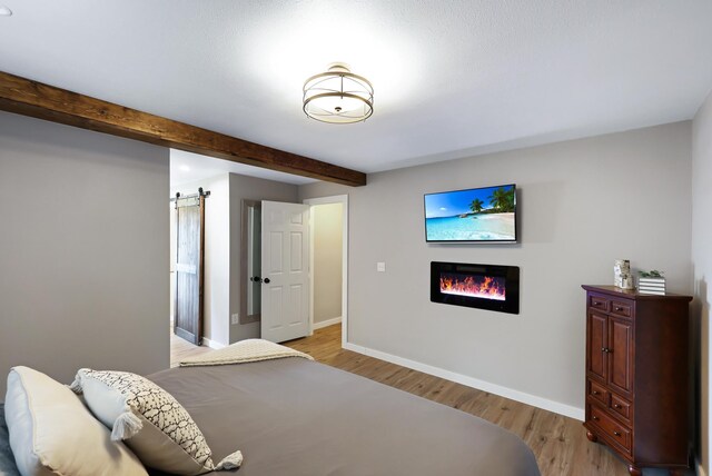 bedroom featuring a barn door, baseboards, light wood-style floors, beam ceiling, and a glass covered fireplace