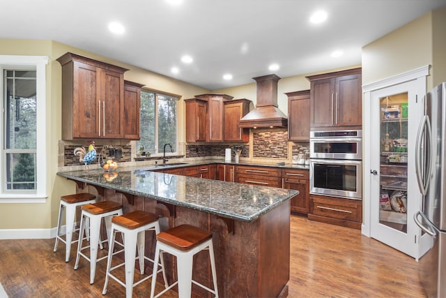 kitchen featuring a sink, stainless steel appliances, dark stone counters, a peninsula, and custom exhaust hood