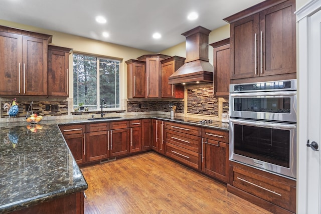 kitchen featuring light wood-type flooring, custom range hood, a sink, backsplash, and stainless steel double oven