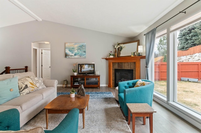 living room featuring baseboards, light wood-style flooring, vaulted ceiling with beams, arched walkways, and a tiled fireplace