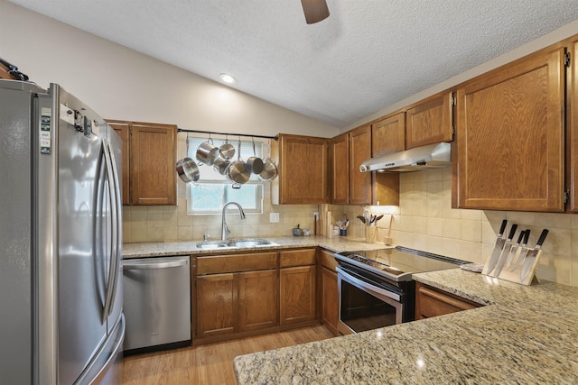 kitchen with brown cabinets, under cabinet range hood, a sink, appliances with stainless steel finishes, and vaulted ceiling