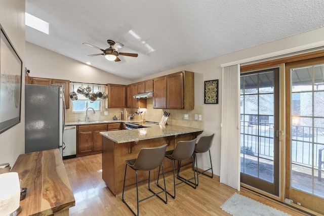 kitchen featuring under cabinet range hood, appliances with stainless steel finishes, a peninsula, brown cabinetry, and a sink