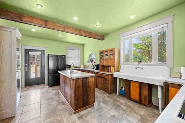 kitchen featuring an island with sink, light countertops, beam ceiling, freestanding refrigerator, and a sink