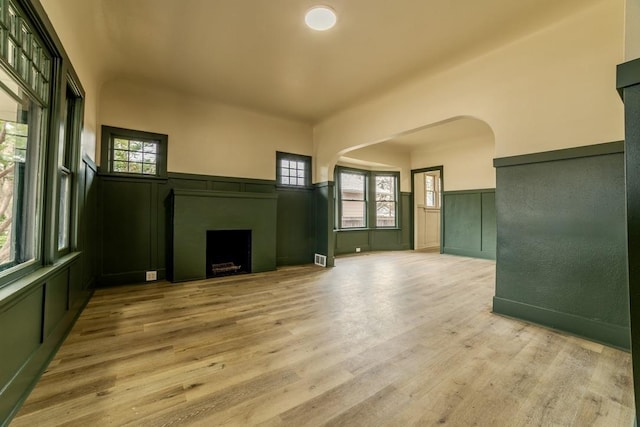 unfurnished living room with a wainscoted wall, light wood-style flooring, a fireplace, arched walkways, and a decorative wall