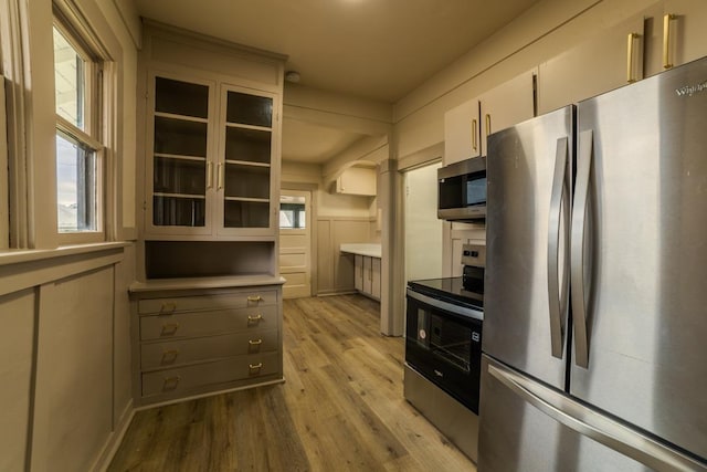 kitchen with light wood finished floors, stainless steel appliances, a decorative wall, glass insert cabinets, and light countertops