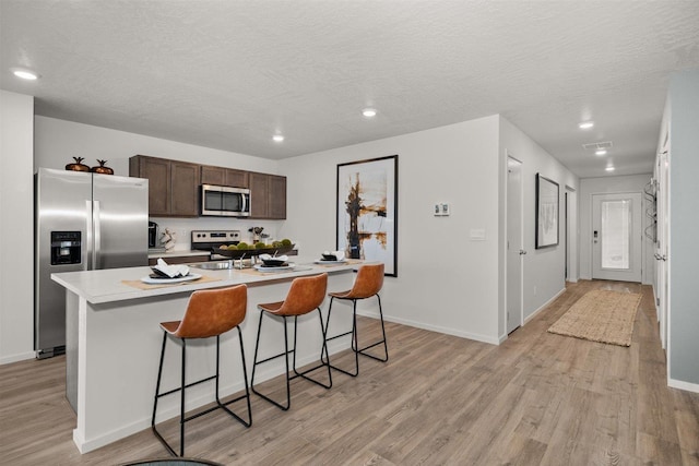 kitchen with a kitchen island with sink, light countertops, dark brown cabinets, appliances with stainless steel finishes, and light wood-type flooring