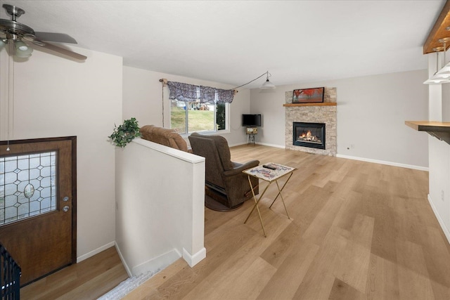 living area featuring light wood finished floors, a stone fireplace, a ceiling fan, and baseboards