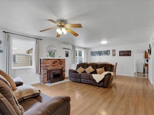 living area with light wood finished floors, a wall mounted AC, arched walkways, ceiling fan, and a brick fireplace