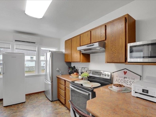 kitchen with a wall mounted air conditioner, brown cabinets, under cabinet range hood, appliances with stainless steel finishes, and light countertops