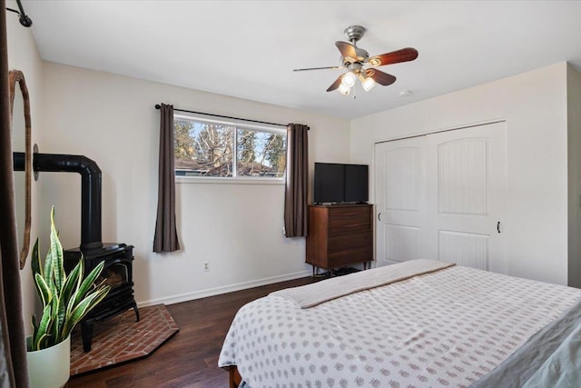 bedroom featuring a ceiling fan, dark wood-style floors, a closet, baseboards, and a wood stove