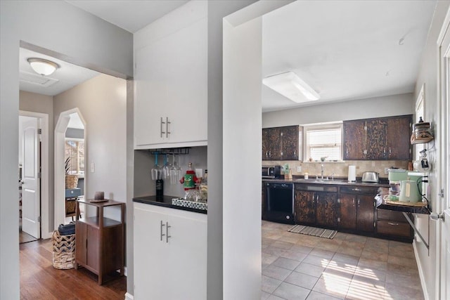 kitchen with arched walkways, black dishwasher, dark brown cabinets, dark countertops, and backsplash