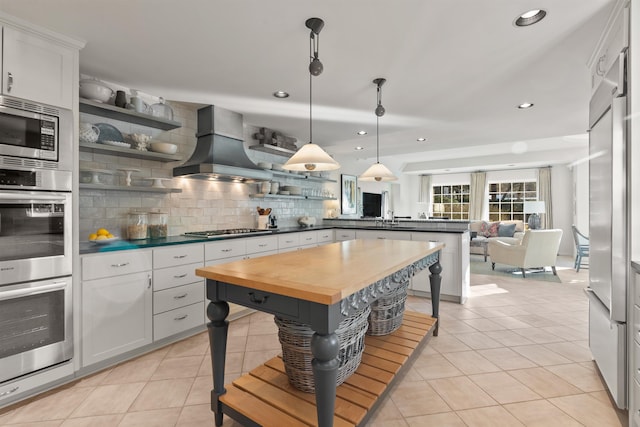 kitchen with custom range hood, open shelves, a peninsula, butcher block counters, and built in appliances