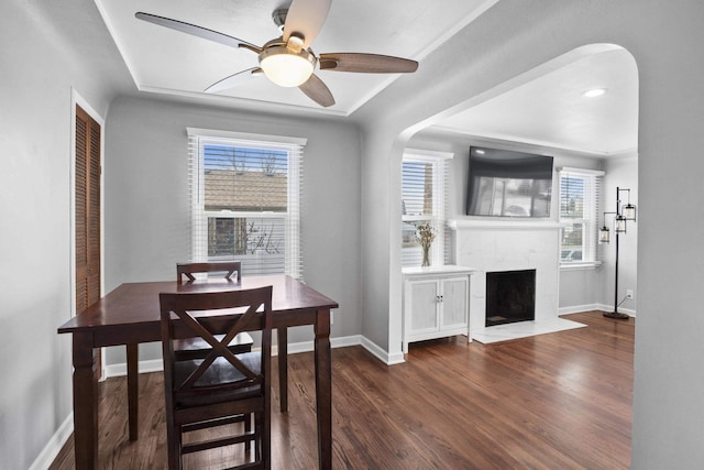 dining room featuring wood finished floors, baseboards, a premium fireplace, arched walkways, and ceiling fan