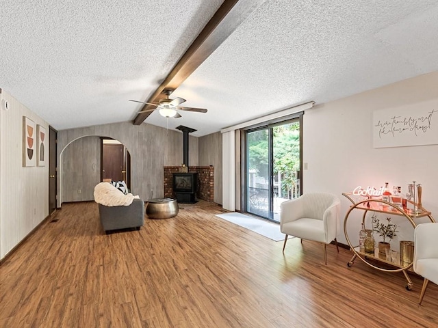 living area featuring wood finished floors, a wood stove, vaulted ceiling with beams, arched walkways, and a textured ceiling