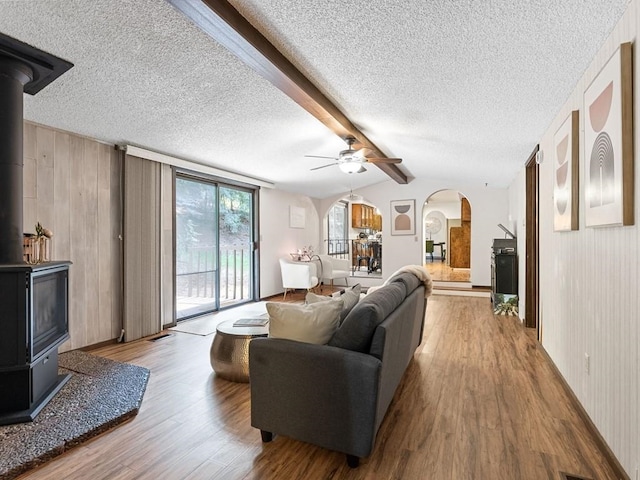 living room with vaulted ceiling with beams, a wood stove, wood finished floors, arched walkways, and a textured ceiling