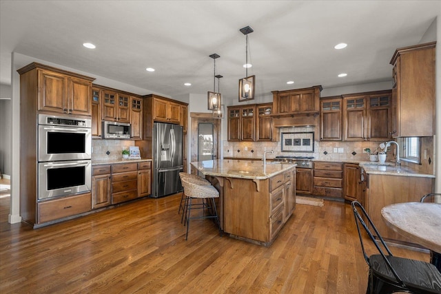 kitchen with light stone countertops, an island with sink, dark wood-style floors, appliances with stainless steel finishes, and brown cabinetry