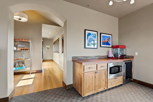 kitchen with stainless steel microwave, light brown cabinets, wine cooler, wood finished floors, and arched walkways