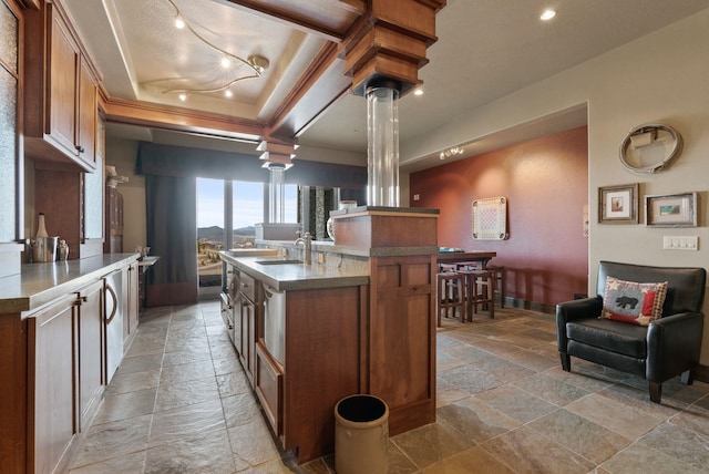 kitchen featuring an island with sink, a sink, stone tile flooring, brown cabinetry, and a raised ceiling