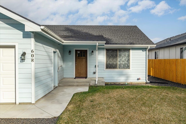 doorway to property featuring a yard, roof with shingles, an attached garage, and fence