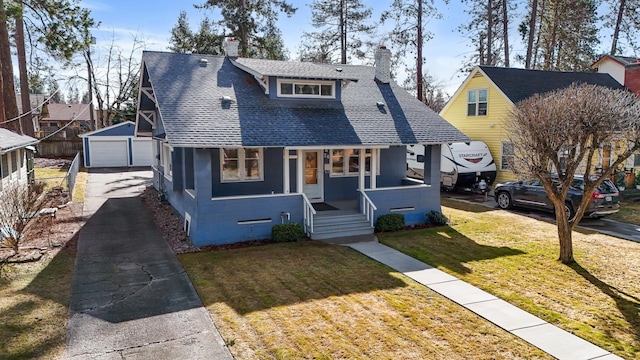 bungalow-style house with an outbuilding, covered porch, roof with shingles, a front yard, and a chimney