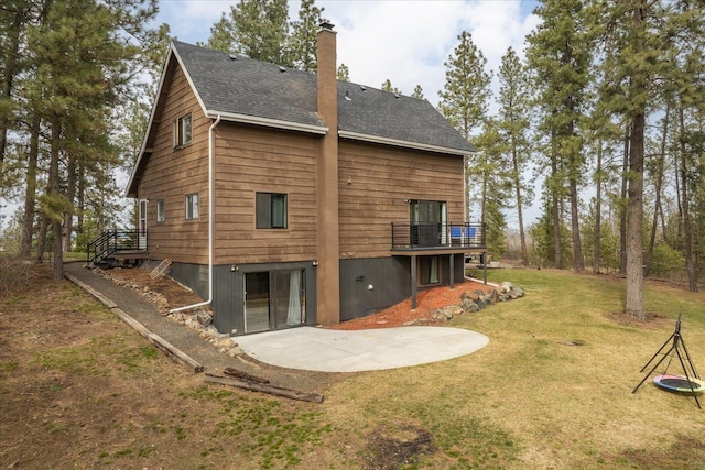 rear view of house with a lawn, a shingled roof, a wooden deck, a chimney, and a patio area