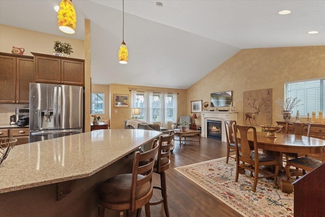 kitchen with light stone countertops, vaulted ceiling, stainless steel refrigerator with ice dispenser, dark wood-style floors, and a glass covered fireplace