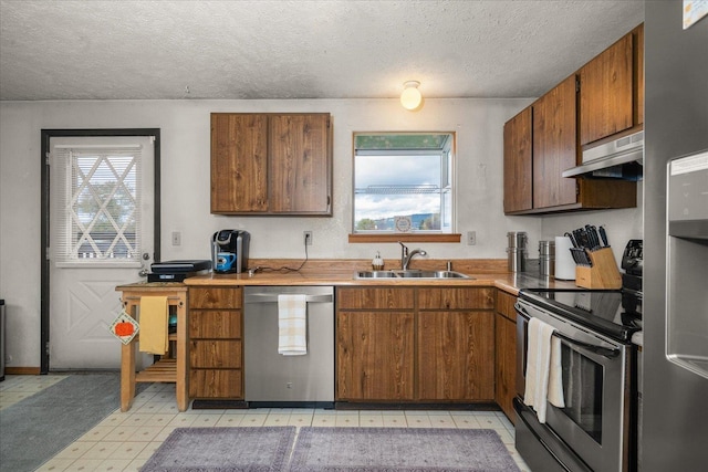 kitchen with under cabinet range hood, a sink, a textured ceiling, appliances with stainless steel finishes, and light floors