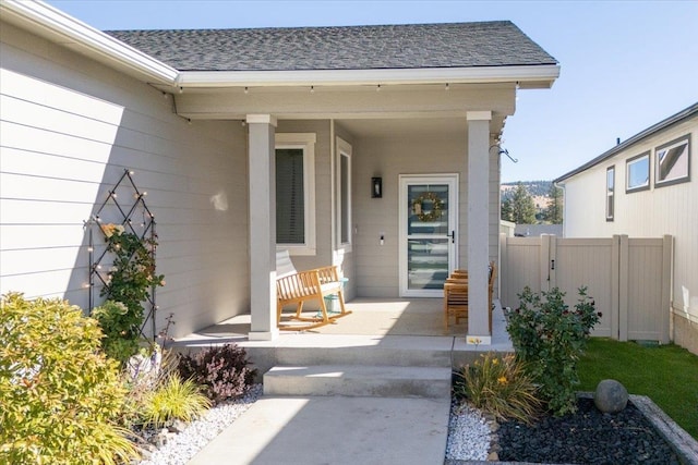 view of exterior entry with covered porch, roof with shingles, and fence