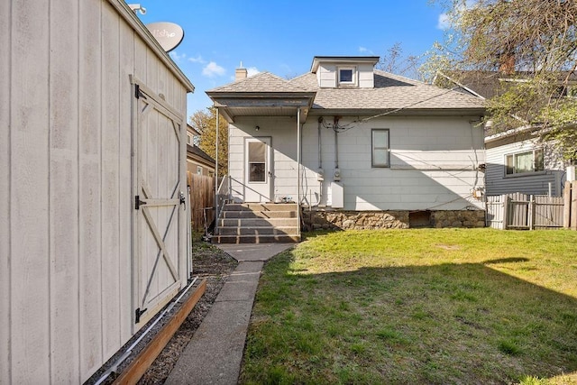 rear view of house featuring fence, an outbuilding, a yard, and a shingled roof