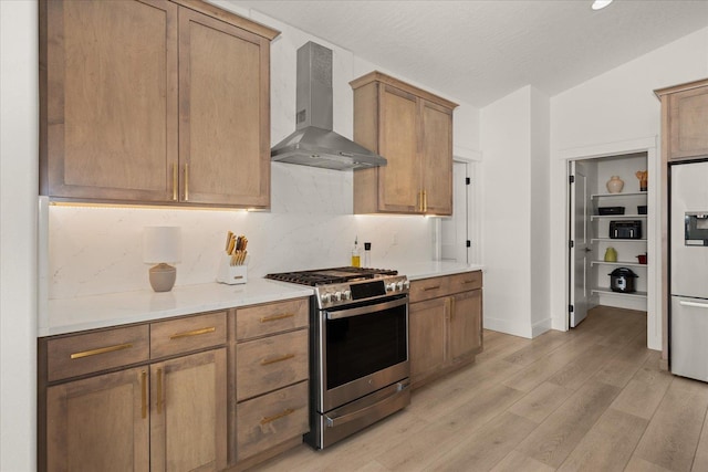 kitchen featuring wall chimney range hood, light wood-style floors, backsplash, refrigerator with ice dispenser, and stainless steel gas range oven