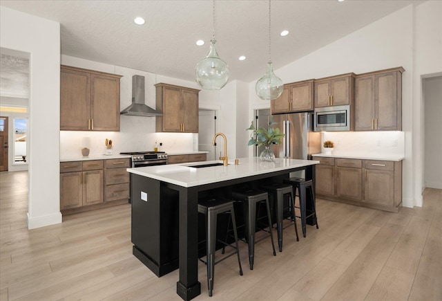 kitchen featuring wall chimney exhaust hood, stainless steel appliances, a kitchen island with sink, a sink, and tasteful backsplash
