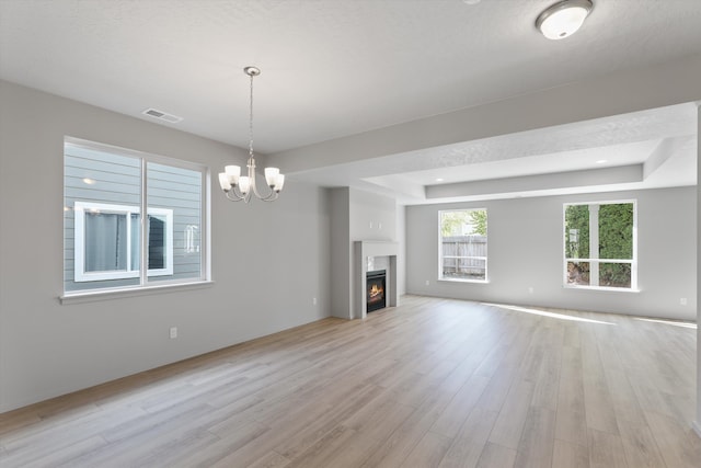 unfurnished living room with a tray ceiling, a warm lit fireplace, light wood-type flooring, visible vents, and an inviting chandelier