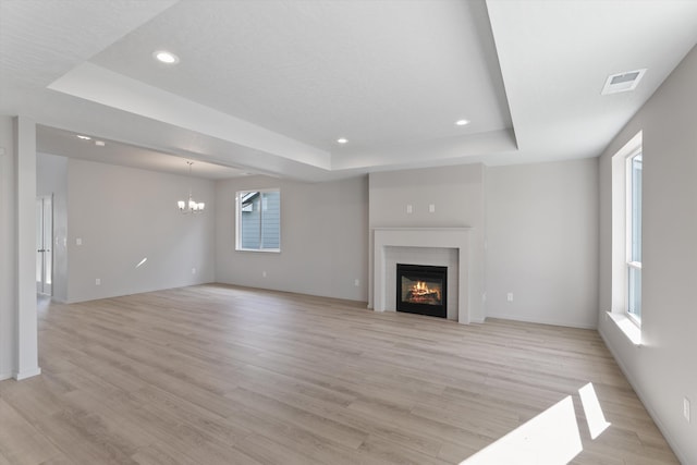 unfurnished living room featuring a tray ceiling, a notable chandelier, a warm lit fireplace, light wood-style flooring, and recessed lighting