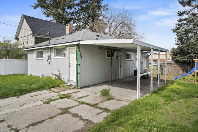 back of house featuring a chimney, fence, a shingled roof, an attached carport, and a lawn